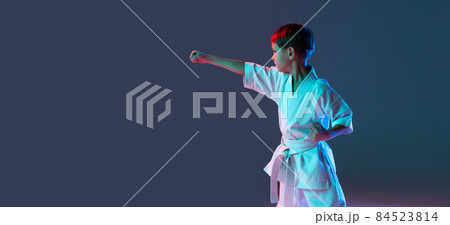 Cropped portrait of boy, karateka training front stance pose isolated over isolated over gradient blue purple background in neon. Flyer 84523814