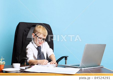 One little boy, child businessman sitting at office table isolated on blue studio background. Concept of smart childhood One little boy, child businessman sitting at office table isolated on blue studio background. Concept of smart childhood 84524291