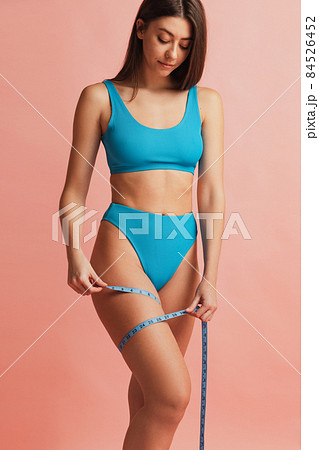 Cropped portrait of slim female model with well kept skin in blue color underwear using centimeter isolated over pink studio background. 84526452