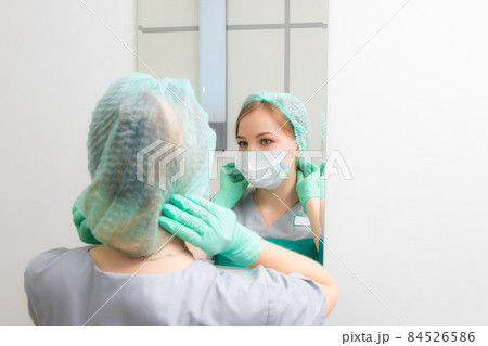 young woman doctor in gloves looks in the mirror and adjusts the cap on her head young woman doctor in gloves looks in the mirror and adjusts the cap on her head 84526586