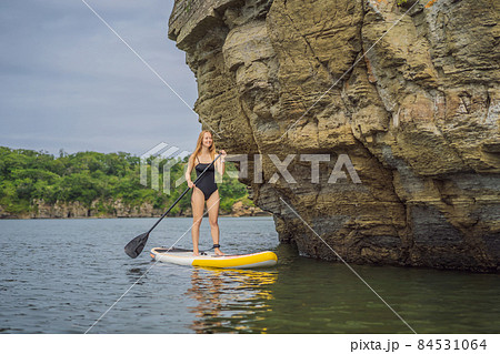 Young women Having Fun Stand Up Paddling in the sea. SUP. Red hair girl Training on Paddle Board near the rocks Young women Having Fun Stand Up Paddling in the sea. SUP. Red hair girl Training on Paddle Board near the rocks 84531064