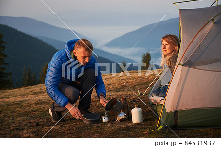 Camping in the morning. Hiker man sitting squatting and pouring water to make coffee near his wife, who admiring the surrounding beauty at dawn from their tent against misty mountains. Camping in the morning. Hiker man sitting squatting and pouring water to make coffee near his wife, who admiring the surrounding beauty at dawn from their tent against misty mountains. 84531937