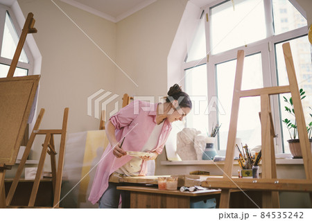 Cute woman paints on canvas in an art workshop. Artist creating picture. Art school or studio. Work with paints, brushes and easel. Hobby and leisure concept. Woman painter at workspace Cute woman paints on canvas in an art workshop. Artist creating picture. Art school or studio. Work with paints, brushes and easel. Hobby and leisure concept. Woman painter at workspace 84535242