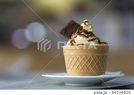White plate with ice cream dessert in wafer cup with chocolate cookies and creative decoration topping on blurred colorful interior background. White plate with ice cream dessert in wafer cup with chocolate cookies and creative decoration topping on blurred colorful interior background. 84539829