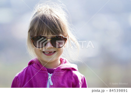 Portrait of cute pretty little blond preschool girl in pink sweater and dark sunglasses smiling happily in camera with funny child toothless smile against blurred light blue background outdoors. Portrait of cute pretty little blond preschool girl in pink sweater and dark sunglasses smiling happily in camera with funny child toothless smile against blurred light blue background outdoors. 84539891