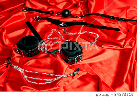Leather collar with metal chain leash, gag ball, handcuffs, nipple clamps on a red satin sheet. Leather collar with metal chain leash, gag ball, handcuffs, nipple clamps on a red satin sheet. 84573219