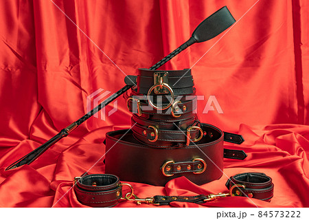 Adult sex games. BDSM items. Leather straps handcuffs, collar, belt and whip on a red satin sheet. Adult sex games. BDSM items. Leather straps handcuffs, collar, belt and whip on a red satin sheet. 84573222