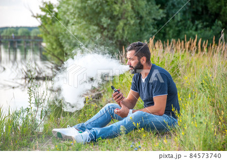 Handsome smoker blowing a lot of smoke using vape electronic cigarette. Hybrid cigarette. Handsome smoker blowing a lot of smoke using vape electronic cigarette. Hybrid cigarette. 84573740
