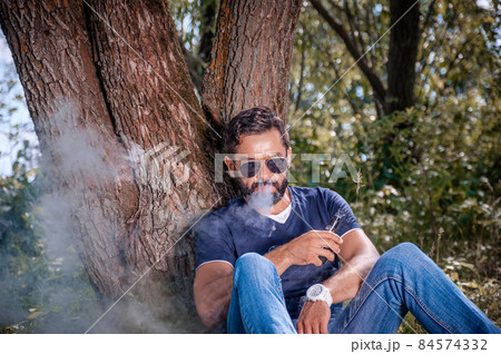 Bearded smoker enjoying an electronic cigarette in the calm outdoors. Hybrid cigarette. Bearded smoker enjoying an electronic cigarette in the calm outdoors. Hybrid cigarette. 84574332