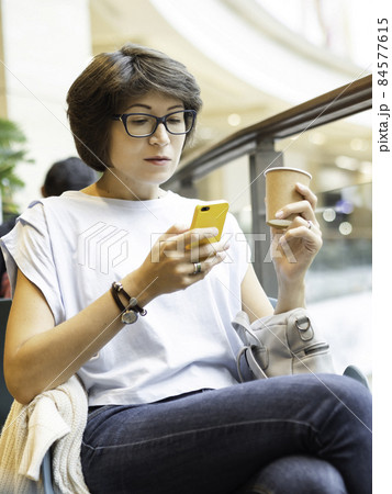 Woman is texting or internet surfing at smartphone and drinking coffee at shopping mall. Coffee break at store. 84577615
