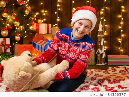 Child girl posing in new year or christmas decoration. Festive lights and lots of gifts, an elegant Christmas tree with toys. The girl is wearing a red sweater and a Santa hat, she plaing with bear 84596577