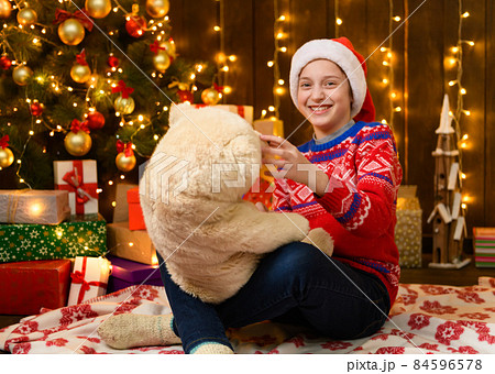Child girl posing in new year or christmas decoration. Festive lights and lots of gifts, an elegant Christmas tree with toys. The girl is wearing a red sweater and a Santa hat, she plaing with bear 84596578