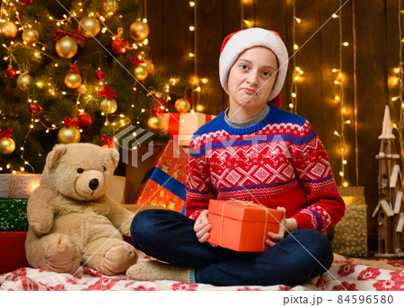 Child girl posing in new year or christmas decoration. Holiday lights and lots of gifts, an elegant Christmas tree with toys. The girl is wearing a red sweater and a Santa hat, she opening a gift box 84596580