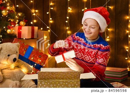 Child girl posing in new year or christmas decoration, wearing a red sweater and a Santa hat, she opening a gift box and having fun. Holiday lights and lots of gifts, Christmas tree with toys. 84596598