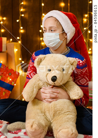 Child girl as Santa helper posing in new year decoration. Wearing a protective face mask against viruses, coronaviruses. She's playing with a toy bear. Holiday lights and lots of gifts, an elegant 84596615