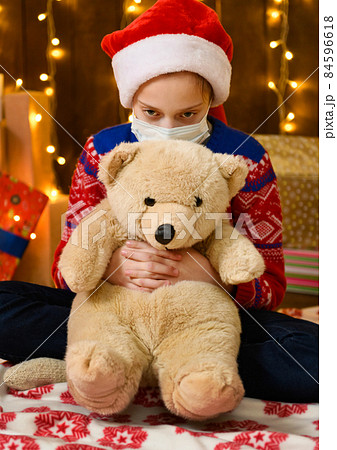 Child girl as Santa helper posing in new year decoration. Wearing a protective face mask against viruses, coronaviruses. She's playing with a toy bear. Holiday lights and lots of gifts, an elegant 84596618