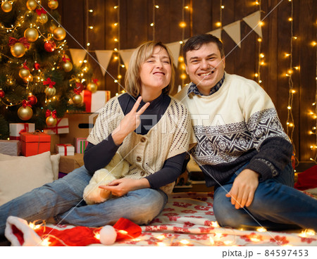 Portrait of a couple in New Year decoration. they show a heavy metal gesture - goat horns. Festive lights, gifts and a Christmas tree decorated with toys. 84597453