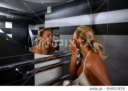 Charming young Caucasian blonde woman in swimsuit applying moisturizer, performing smoothing rejuvenating facial massage looking at her reflection in bathroom mirror 84601859