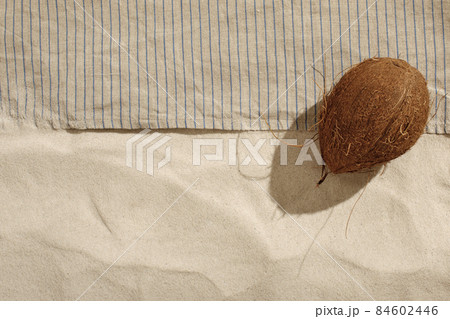 Striped linen beach towel with fringes and coconut on sandy beach with shadows from palm tree. Relaxation and tropical summer holidays concept Striped linen beach towel with fringes and coconut on sandy beach with shadows from palm tree. Relaxation and tropical summer holidays concept 84602446