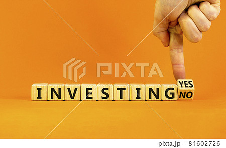 Investing yes or no symbol. Businessman turns a wooden cube and change words 'investing no' to 'investing yes'. Beautiful orange background. Business and investing yes or no concept, copy space. 84602726
