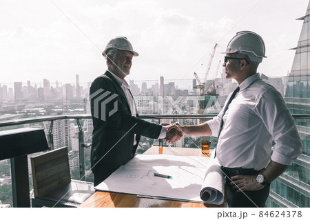 Two engineering working together in their new project. They are wearing suit , tie and white hard hat. They are looking at their design on the paper on the wooden table Two engineering working together in their new project. They are wearing suit , tie and white hard hat. They are looking at their design on the paper on the wooden table 84624378