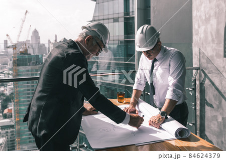 Two engineering working together in their new project. They are wearing suit , tie and white hard hat. They are looking at their design on the paper on the wooden table Two engineering working together in their new project. They are wearing suit , tie and white hard hat. They are looking at their design on the paper on the wooden table 84624379