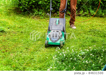 Man cutting green grass with lawn mower in backyard. Gardening country lifestyle background. Beautiful view on fresh green grass lawn in sunlight, garden landscape in spring or summer season Man cutting green grass with lawn mower in backyard. Gardening country lifestyle background. Beautiful view on fresh green grass lawn in sunlight, garden landscape in spring or summer season 84631500