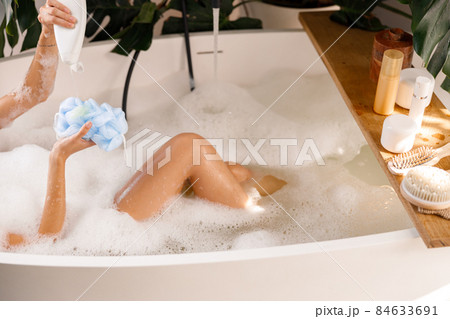 Female hands pouring shower gel from bottle onto loofah sponge. Wooden shelf with body care products over bubble filled bathtub 84633691