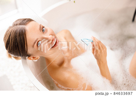 Happy young woman smiling at camera, holding disposable shaving razor for shaving her legs in the bath with foam 84633697