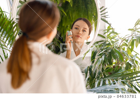 Young woman wearing bathrobe looking at herself in the mirror while using silicone face brush to cleanse skin, standing in bathroom decorated with green plants 84633781