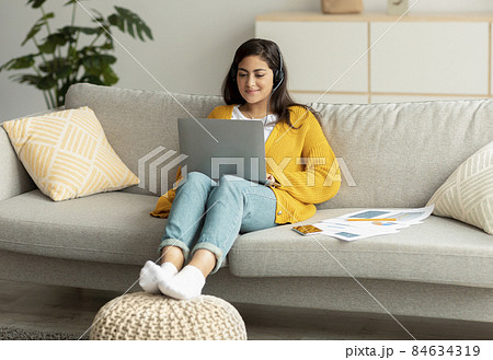 Telecommuting concept. Excited arab woman sitting on sofa and working online on laptop, wearing headset 84634319