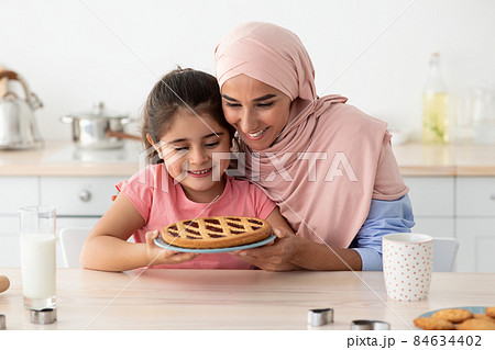 Happy Muslim Mother And Cute Little Daughter Holding Homemade Pie In Kitchen Happy Muslim Mother And Cute Little Daughter Holding Homemade Pie In Kitchen 84634402