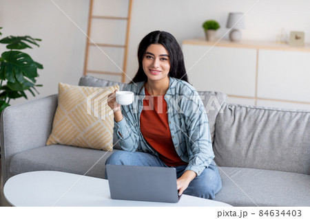 Lovely young Arab lady using laptop, studying or working online, having remote business meeting, drinking coffee at home 84634403