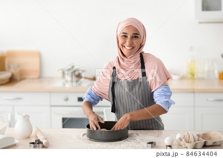 Happy Muslim Woman Pressing Dough Into Mold While Baking In Kitchen Happy Muslim Woman Pressing Dough Into Mold While Baking In Kitchen 84634404
