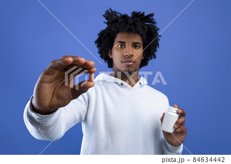 Black teenager holding jar of tablets and showing one pill over blue studio background, selective focus Black teenager holding jar of tablets and showing one pill over blue studio background, selective focus 84634442