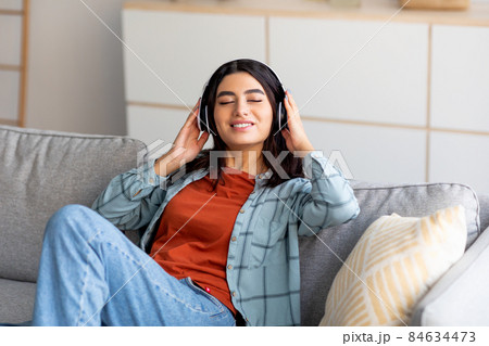 Portrait of young Arab woman in headphones relaxing on couch and listening to music with closed eyes at home Portrait of young Arab woman in headphones relaxing on couch and listening to music with closed eyes at home 84634473