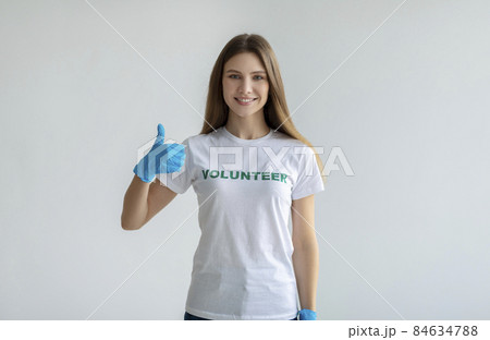 I like volunteering. Positive lady in white volunteer t-shirt showing thumb up, wearing blue gloves, light background 84634788
