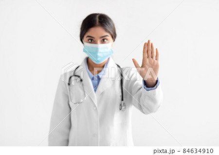 Serious young indian woman doctor in coat and protective mask show stop hand gesture 84634910