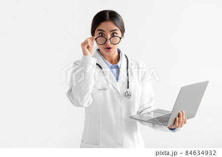 Surprised young hindu female doctor in white coat takes off glasses and looks at camera, works at laptop Surprised young hindu female doctor in white coat takes off glasses and looks at camera, works at laptop 84634932