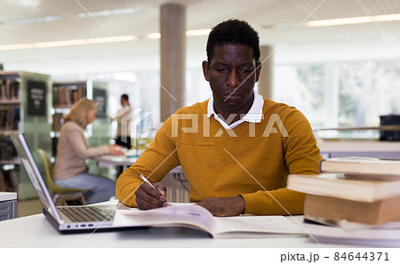 Efficient man preparing for exam in public library, reading book and using laptop for searching information 84644371