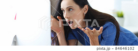 Shocked woman sits at table and looks at computer screen eyes widening in horror 84646102