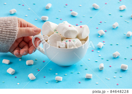 A woman's hand holds a cup of hot chocolate, lavishly decorated with marshmallows on a blue background. 84648228