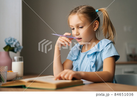 Close-up of thinking pretty elementary child school girl doing homework and holding pen against mouth sitting at home table. 84651036