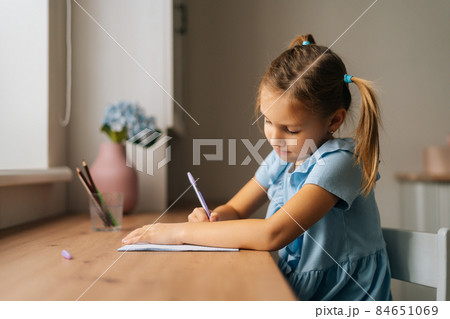 Side view of focused primary little child girl learning writing doing homework sitting at home table by window. 84651069