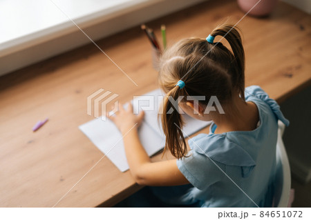 High-angle view of unrecognizable little child girl learning writing doing homework sitting at home table by window. High-angle view of unrecognizable little child girl learning writing doing homework sitting at home table by window. 84651072