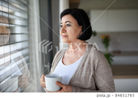 Sad senior woman standing and looking out through window indoors at home. Sad senior woman standing and looking out through window indoors at home. 84651765
