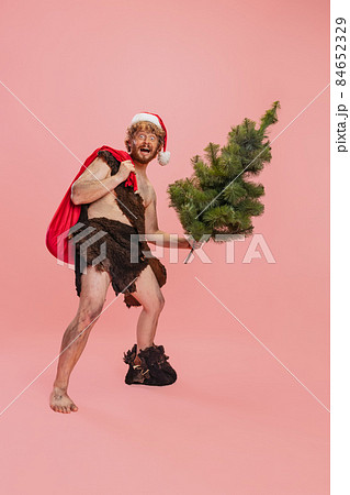 Full-length portrait of joyful man, neanderthal wearing red holiday hat, holding Christmas tree and log isolated over pink background. Merry holidays 84652329