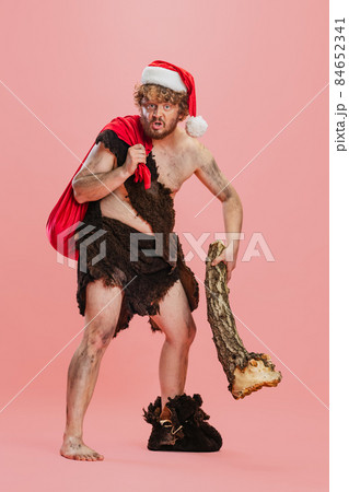 Full-length portrait of man in character neanderthal, primitive person holding log and Christmas presents bag isolated over pink background Full-length portrait of man in character neanderthal, primitive person holding log and Christmas presents bag isolated over pink background 84652341