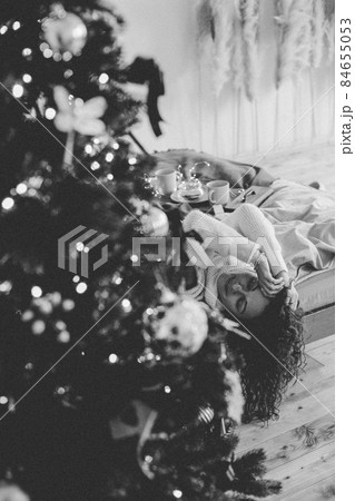 A young woman in cozy home clothes poses on a bed in a loft interior near a Christmas tree. A young woman in cozy home clothes poses on a bed in a loft interior near a Christmas tree. 84655053