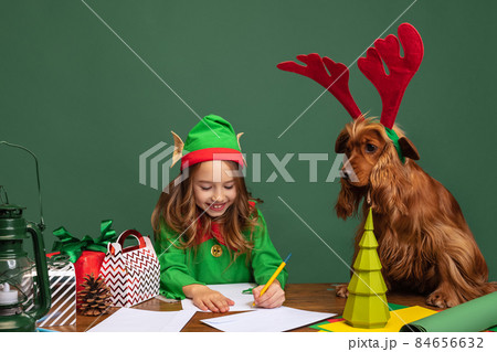 Little beautiful kid dressed like funny gnome or elf sitting at table with her funny dog and writing letter to Santa Claus 84656632